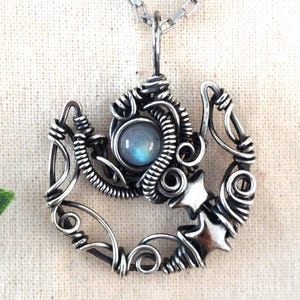 May include: A silver pendant necklace with a wire-wrapped design. The pendant features a central, round, pale blue stone, surrounded by detailed wirework and star-shaped accents. The necklace hangs from a fine silver chain.