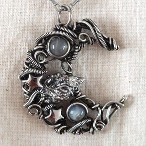 May include: A silver crescent moon pendant necklace. The pendant has a wire-wrapped design with two round, grey gemstones and a detailed centre with a star and leaf motif. The necklace hangs on a delicate chain.