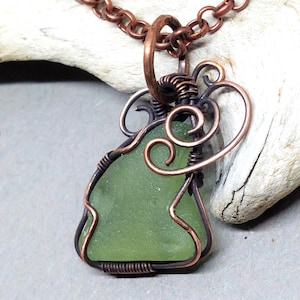 May include: A handcrafted necklace featuring a sea glass pendant in a light green hue, intricately wrapped with copper wire. The pendant hangs from a copper chain, creating a rustic and artistic aesthetic. The necklace is displayed on a piece of driftwood.