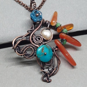 May include: A handcrafted copper wire pendant necklace featuring a cross-shaped design. The pendant is adorned with turquoise, pearl, and orange-red gemstones, along with a blue flower-shaped bead. The necklace chain is a matching copper color.