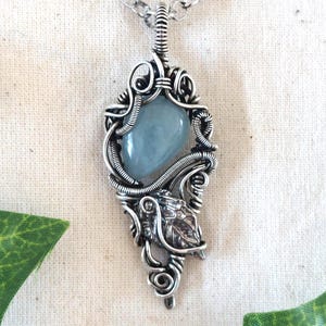 May include: A silver pendant necklace with a light blue gemstone. The pendant has an intricate wire-wrapped design and a leaf accent. The necklace is displayed on a cream-coloured fabric.