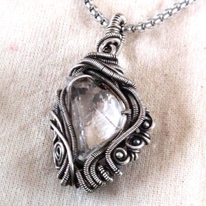 May include: A handcrafted silver pendant necklace featuring a clear, faceted crystal encased in intricate wire wrapping. The pendant hangs from a delicate silver chain, showcasing a unique, artistic design. The crystal is approximately 2.5 cm in length.