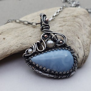 May include: A handcrafted pendant necklace with a pear-shaped, pale blue gemstone. The stone is set in a silver wire frame with decorative swirls, small pearls, and copper accents. The necklace has a silver chain.