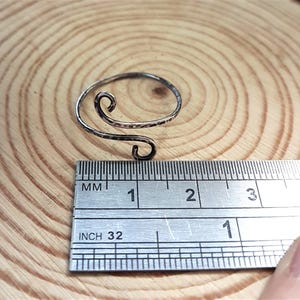 May include: A silver-colored, adjustable ring with a hammered texture and a swirling design. The ring is shown with a ruler, and appears to be approximately 1 inch in diameter.