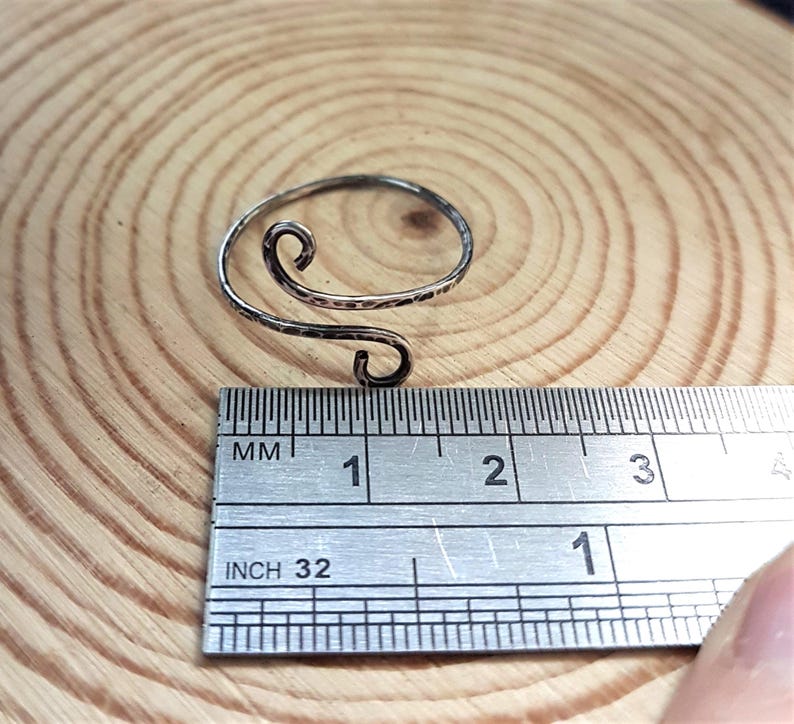 May include: A silver-colored, adjustable ring with a hammered texture and a swirling design. The ring is shown with a ruler, and appears to be approximately 1 inch in diameter.