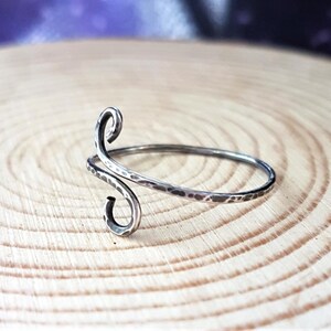 May include: A silver-toned, hammered metal ring with a unique, swirling design. The ring features an open, adjustable band and a decorative, curved element. The ring is displayed on a light brown, wooden surface.