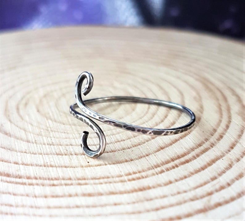 May include: A silver-toned, hammered metal ring with a unique, swirling design. The ring features an open, adjustable band and a decorative, curved element. The ring is displayed on a light brown, wooden surface.
