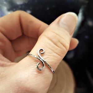 May include: A silver-toned ring with a hammered texture and a stylized, cursive letter design. The ring is worn on a finger, showcasing its intricate, handcrafted details. The ring's design features two swirling elements.
