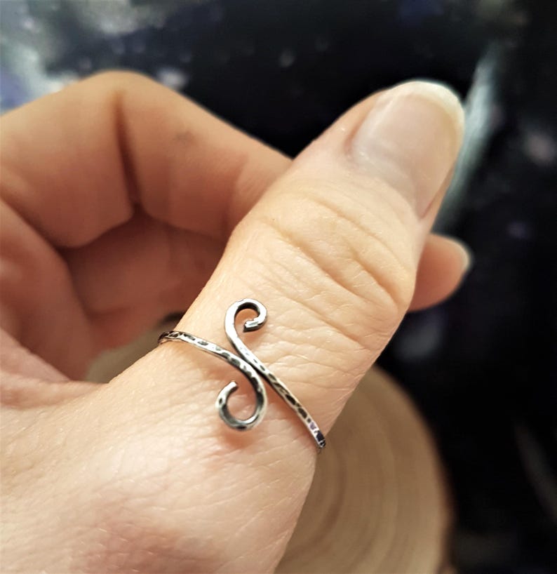 May include: A silver-toned ring with a hammered texture and a stylized, cursive letter design. The ring is worn on a finger, showcasing its intricate, handcrafted details. The ring's design features two swirling elements.