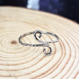 May include: A silver-toned ring with a hammered texture and a unique, open design. The ring features two swirling, curved ends, creating an elegant and artistic look. The ring is displayed on a wooden surface.