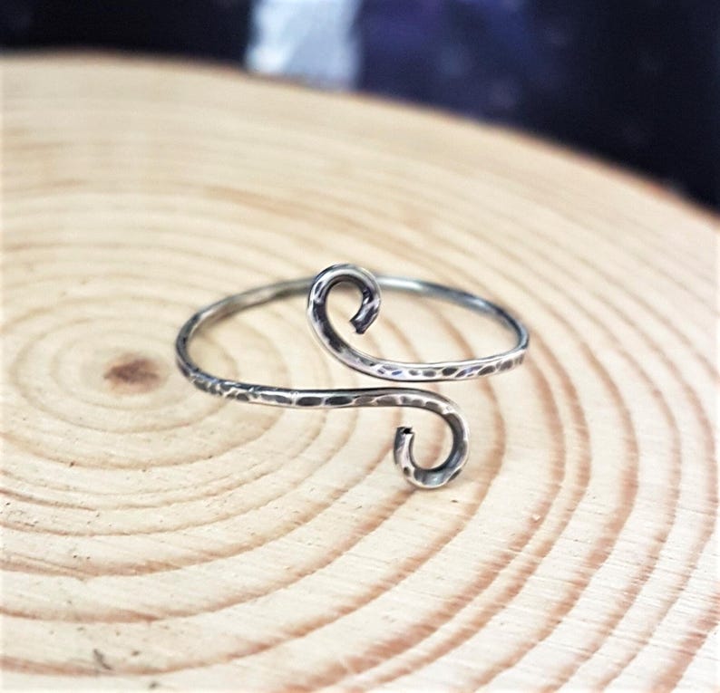 May include: A silver-toned ring with a hammered texture and a unique, open design. The ring features two swirling, curved ends, creating an elegant and artistic look. The ring is displayed on a wooden surface.