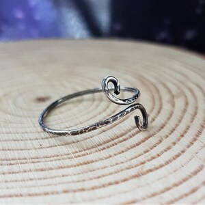 May include: A silver-toned ring with a hammered texture and a unique, swirling design. The ring features two curved ends, creating an open, adjustable style. The ring is displayed on a wooden surface.