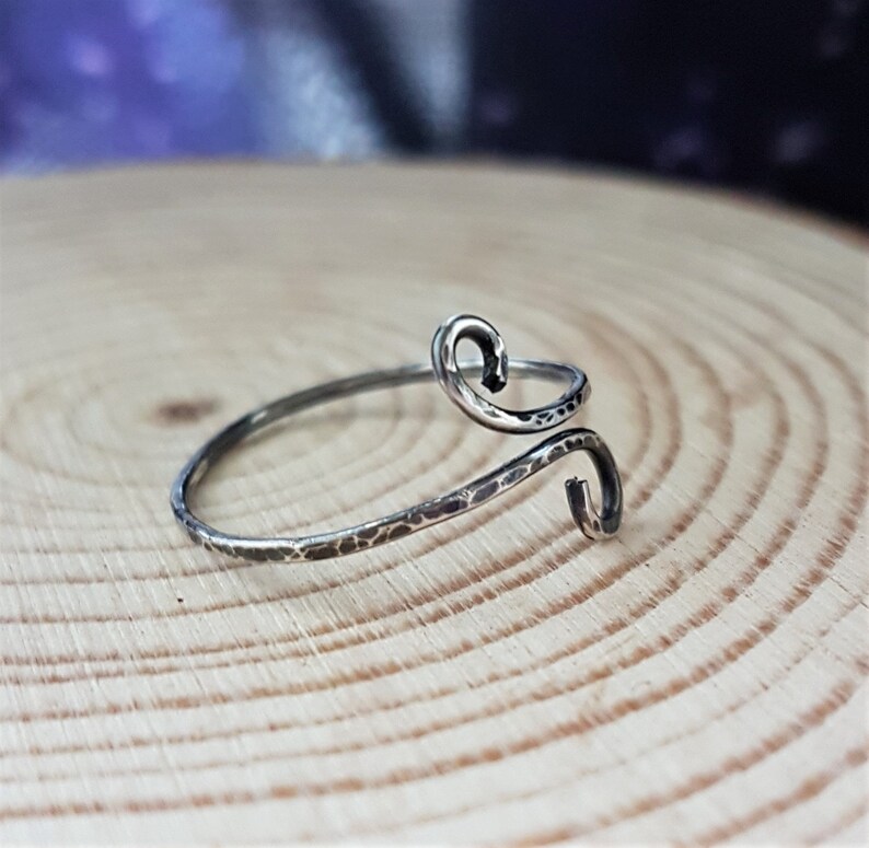May include: A silver-toned ring with a hammered texture and a unique, swirling design. The ring features two curved ends, creating an open, adjustable style. The ring is displayed on a wooden surface.
