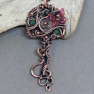 Könnte beinhalten: Kupferfarbener Schlüsselanhänger mit aufwendigem Drahtdesign. Verfügt über einen rosa Schmetterlingsakzent und grüne Edelsteindetails. Der Schlüsselkopf hat ein Spiraldesign. Ein einzigartiges, handgefertigtes Schmuckstück.