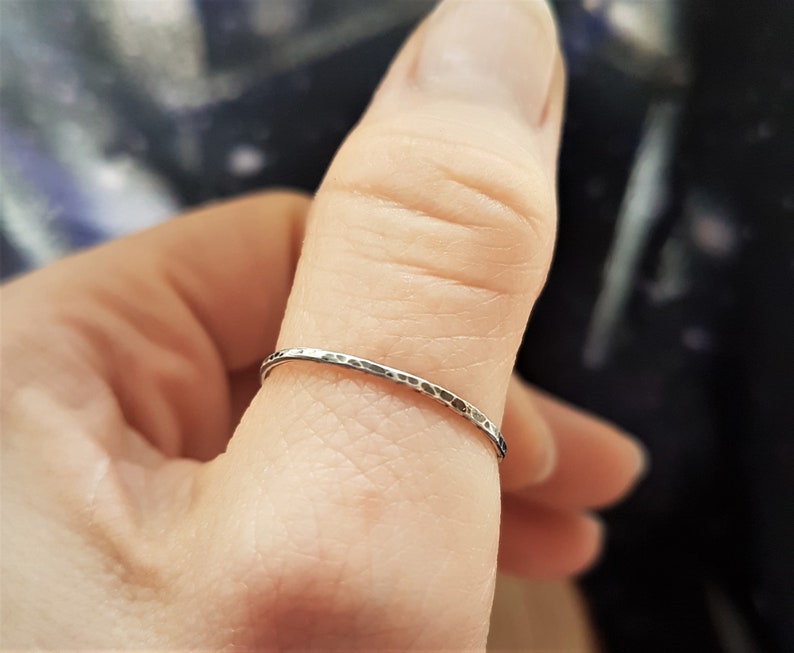 May include: A thin, hammered silver ring. The ring has a textured surface and a simple, minimalist design. The ring is worn on a finger, showcasing its delicate and understated elegance. This piece is suitable for everyday wear.