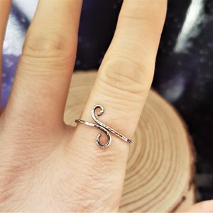 May include: A silver-colored ring with a hammered texture. The ring features a stylized, open design with two curved swirls connected by a straight bar. The ring is worn on a finger.