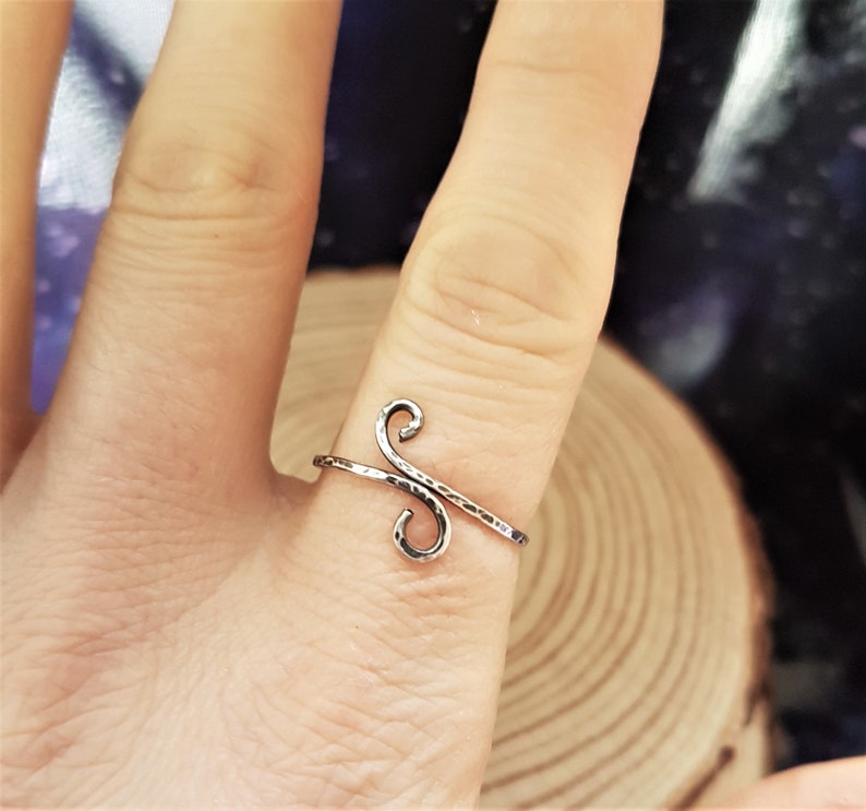 May include: A silver-colored ring with a hammered texture. The ring features a stylized, open design with two curved swirls connected by a straight bar. The ring is worn on a finger.