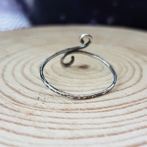 May include: A silver ring with a hammered texture and a swirling, open design. The ring features a delicate, thin band and a decorative spiral at the top. The ring is displayed on a light brown wooden surface.