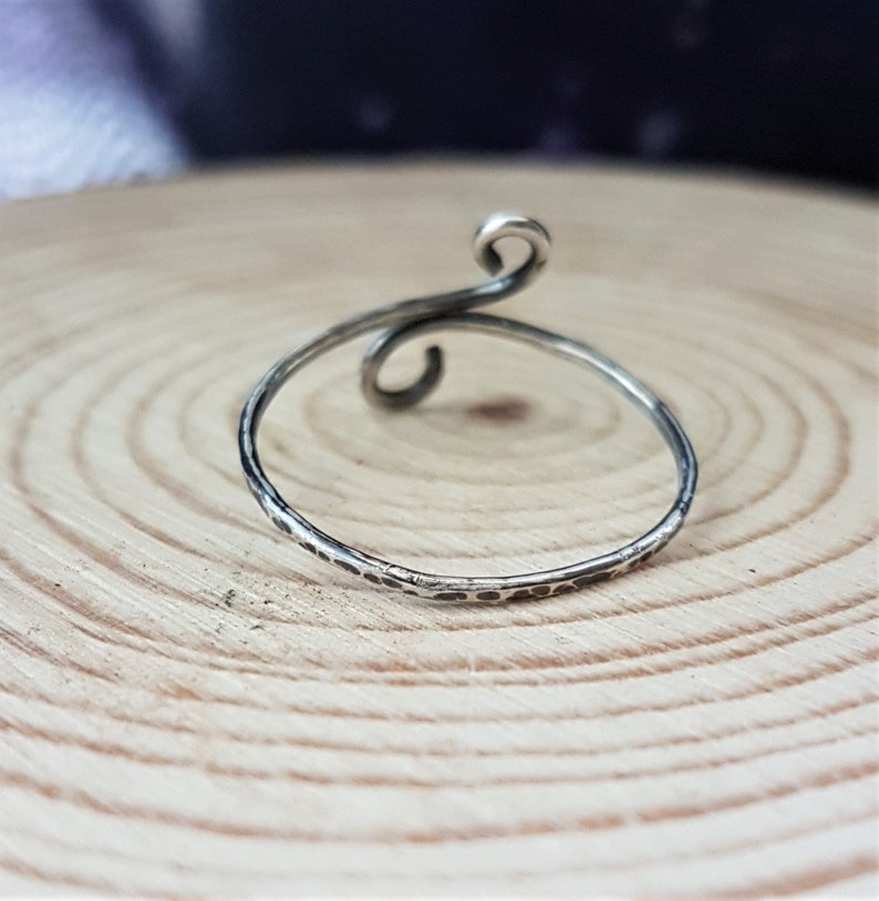 May include: A silver ring with a hammered texture and a swirling, open design. The ring features a delicate, thin band and a decorative spiral at the top. The ring is displayed on a light brown wooden surface.