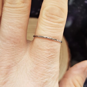 May include: A silver ring with a hammered texture is worn on a finger. The ring is thin and has a slightly irregular surface. The background is blurred, with hints of wood and dark colors.