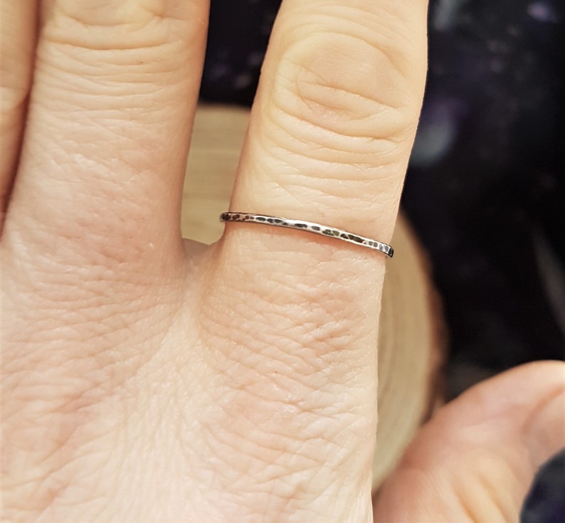 May include: A silver ring with a hammered texture is worn on a finger. The ring is thin and has a slightly irregular surface. The background is blurred, with hints of wood and dark colors.