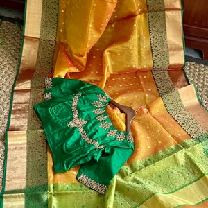 May include: A green silk blouse with gold embroidery, displayed on a yellow and green saree with gold accents.