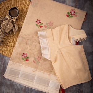 May include: A beige saree and matching blouse set. The saree features floral embroidery in red and pink, with green leaves. The blouse has a round neckline and short sleeves. The saree has a silver border. The text "Mayukha Boutique" is at the bottom.