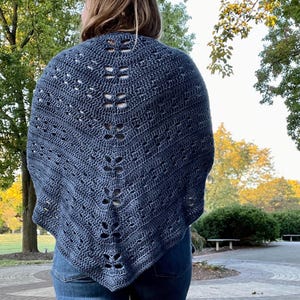 May include: A dark blue crocheted shawl with a dragonfly design. The shawl is worn over a person's shoulders and is paired with blue jeans. The background features trees and a park setting.