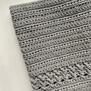 May include: A gray crocheted blanket with a decorative border. The blanket is made with a simple stitch pattern and has a soft, textured feel.
