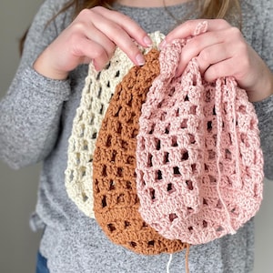 Puede incluir: Tres bolsas de crochet en diferentes colores: crema, marrón y rosa. Las bolsas están hechas con un patrón de punto simple y abierto y tienen una forma cuadrada.