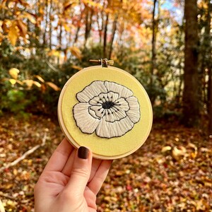 Op de afbeelding: Een witte geborduurde bloem met zwarte accenten op een gele achtergrond. De borduurwerk is in een hoepel en wordt vastgehouden door een hand.