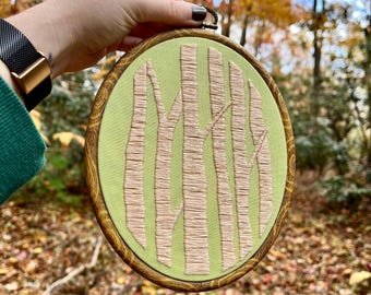 May include: A green embroidery hoop with a beige embroidered design of tree trunks. The hoop is held in a hand against a blurred background of fall foliage.