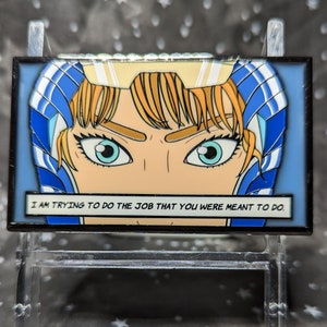 May include: A blue and yellow enamel pin with a cartoon face and the text "I am trying to do the job that you were meant to do."