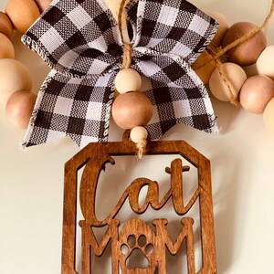 May include: Wooden sign with the text "Cat Mom" and a paw print in the center. The sign is hanging from a string of wooden beads with a black and white checkered bow.