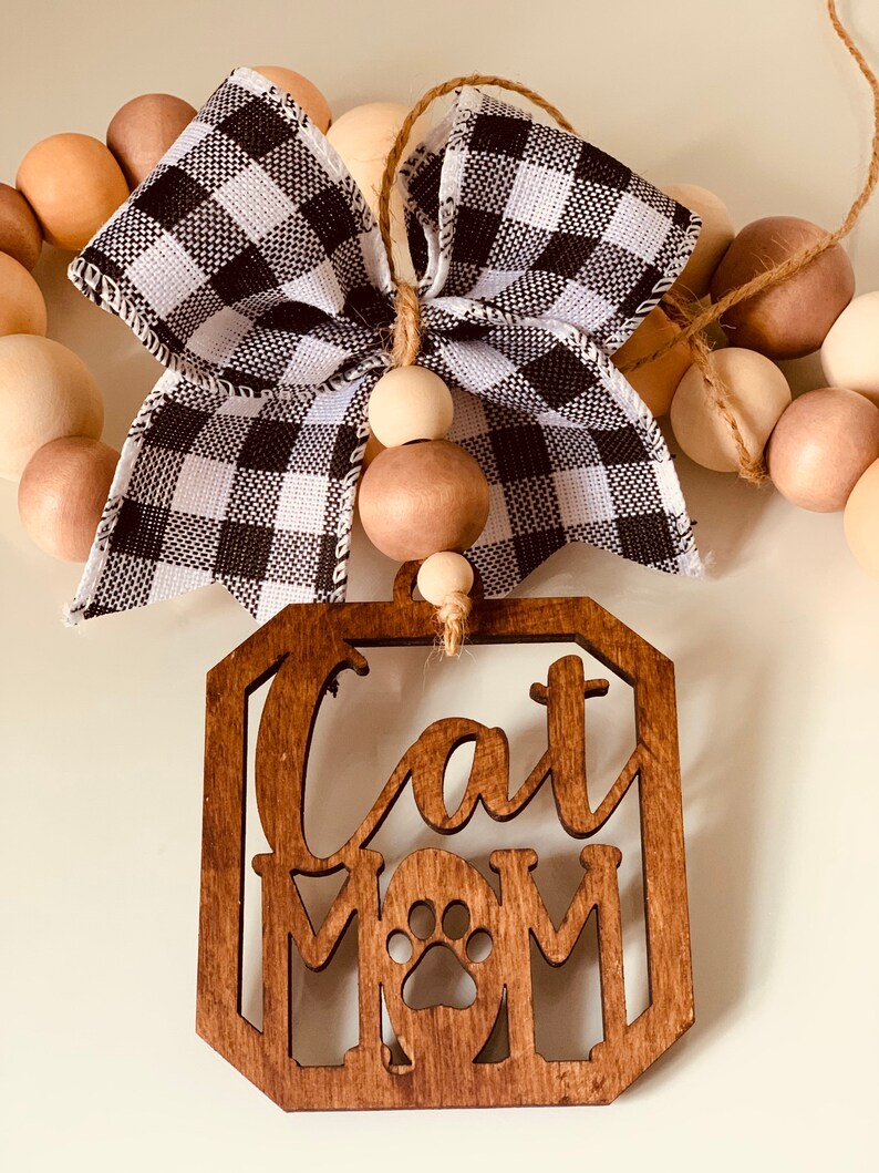 May include: Wooden sign with the text "Cat Mom" and a paw print in the center. The sign is hanging from a string of wooden beads with a black and white checkered bow.