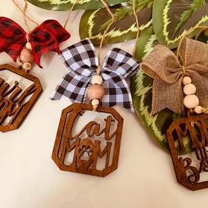 May include: Three wooden ornaments with different designs and bows. The first ornament says "Girl Mom" with a red and black plaid bow. The second ornament says "Cat Mom" with a black and white checkered bow. The third ornament says "Mom of Both" with a brown burlap bow.