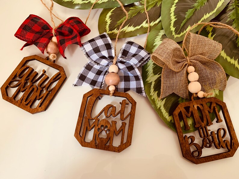 May include: Three wooden ornaments with different designs and bows. The first ornament says "Girl Mom" with a red and black plaid bow. The second ornament says "Cat Mom" with a black and white checkered bow. The third ornament says "Mom of Both" with a brown burlap bow.