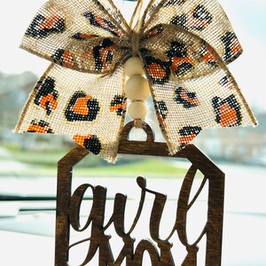 May include: A wooden car charm with the words "Girl Mom" in a decorative script font. The charm is hanging from a beige and black leopard print bow with wooden beads.