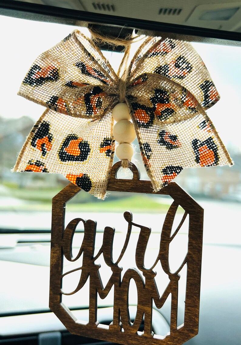 May include: A wooden car charm with the words "Girl Mom" in a decorative script font. The charm is hanging from a beige and black leopard print bow with wooden beads.