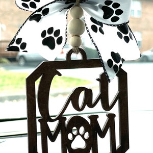 May include: A wooden car charm with the words "Cat Mom" and a paw print in the center. The charm is decorated with a white and black ribbon with paw prints and wooden beads.