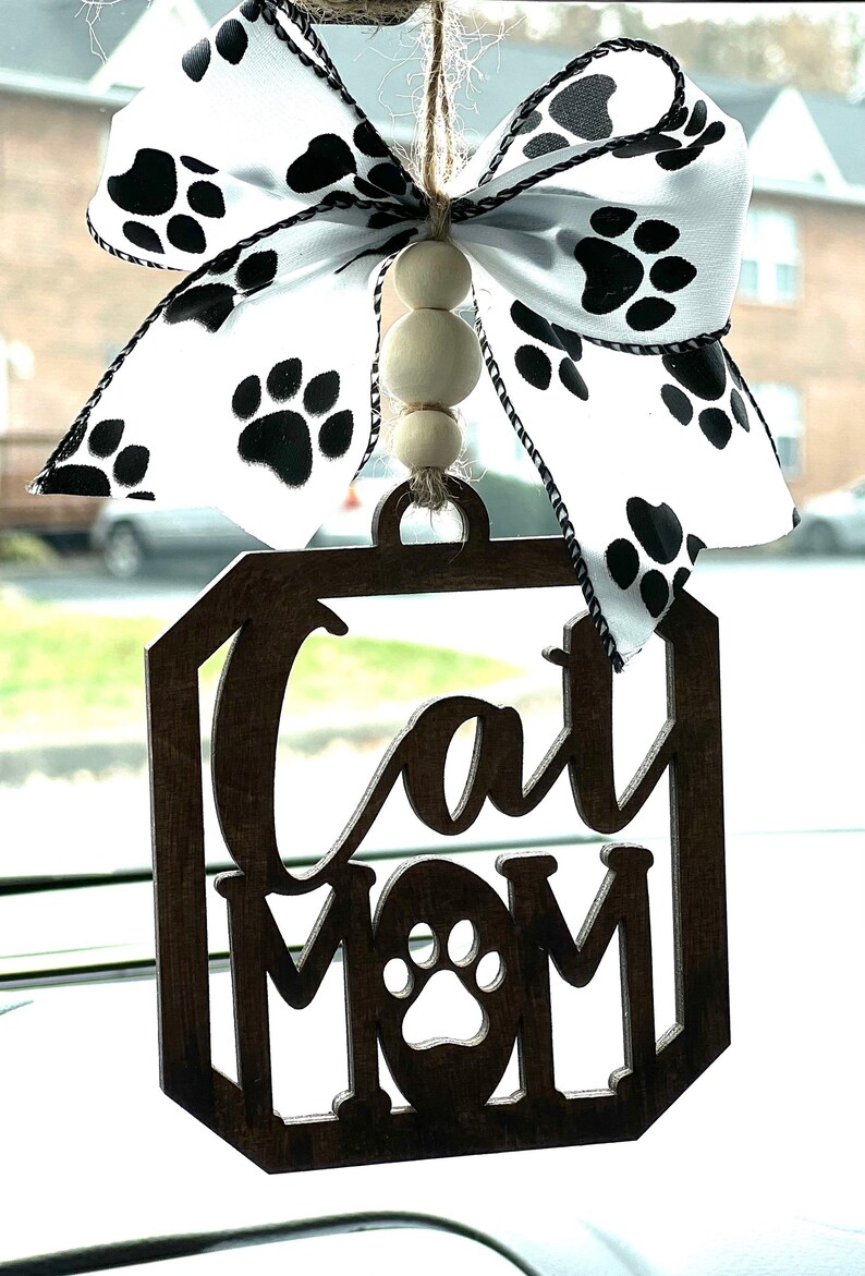 May include: A wooden car charm with the words "Cat Mom" and a paw print in the center. The charm is decorated with a white and black ribbon with paw prints and wooden beads.