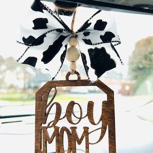 May include: A wooden car charm with the text "boy mom" in a decorative font. The charm is attached to a black and white cow print ribbon with wooden beads.