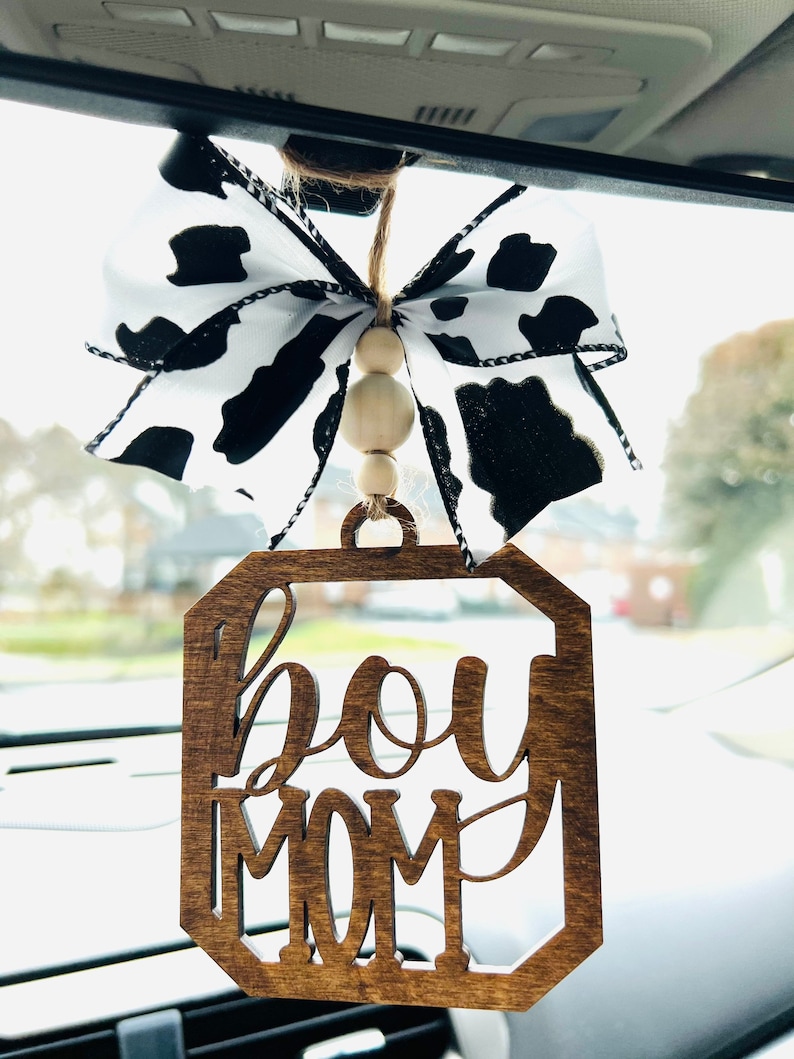 May include: A wooden car charm with the text "boy mom" in a decorative font. The charm is attached to a black and white cow print ribbon with wooden beads.