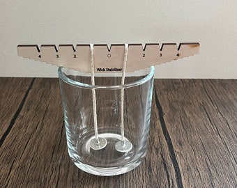 May include: A wooden wick stabilizer with markings from 2 to 4, used to center wicks in candle jars. Two wicks are shown centered in a clear glass jar with metal wick stickers at the bottom.