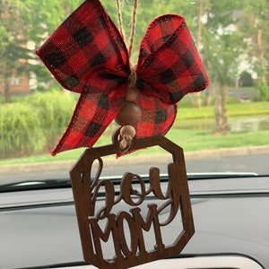 May include: A wooden car charm with the words "Boy Mom" in a decorative font. The charm is hanging from a rearview mirror with a red and black plaid bow.