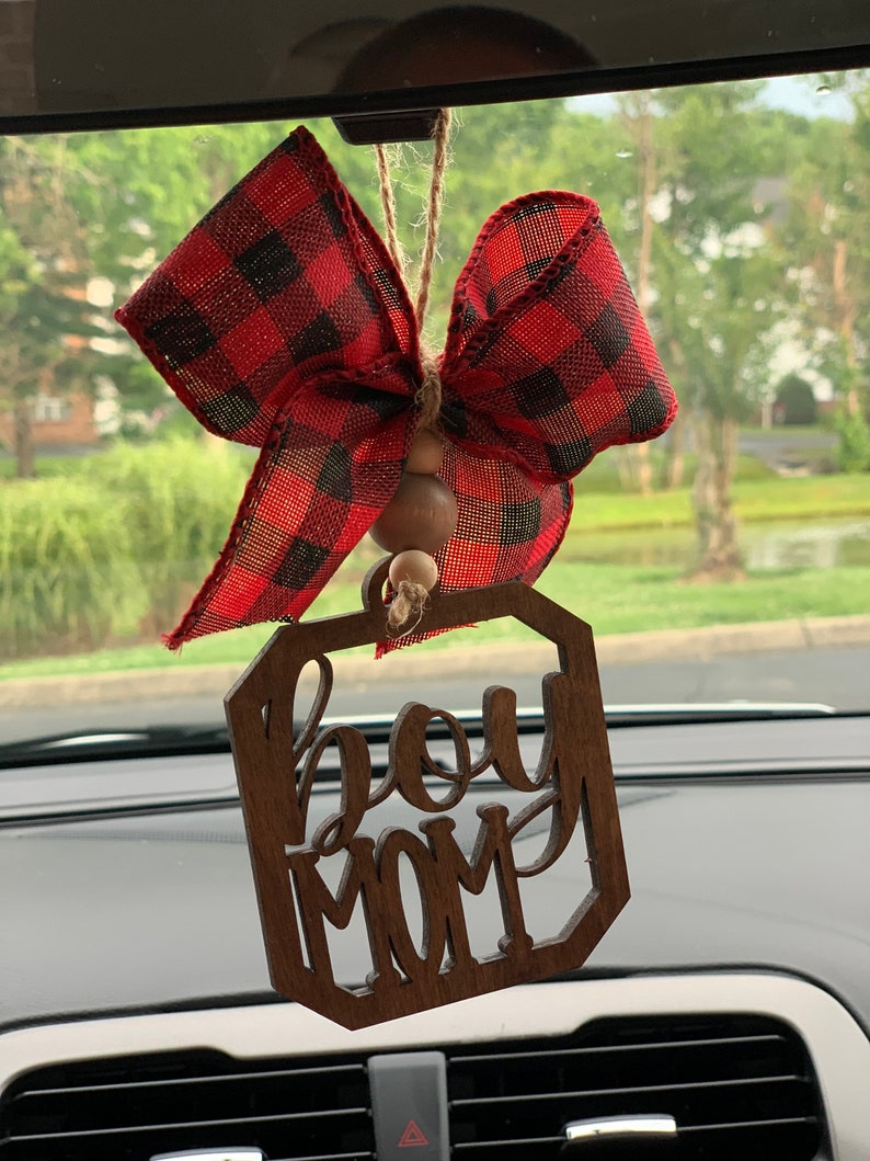 May include: A wooden car charm with the words "Boy Mom" in a decorative font. The charm is hanging from a rearview mirror with a red and black plaid bow.