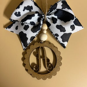May include: A wooden ornament with a scalloped edge and the letter "A" cut out in the center. The ornament is hanging from a black and white cow print ribbon with wooden beads.