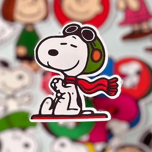May include: A Snoopy sticker in the center, wearing a green pilot helmet with goggles and a red scarf. Snoopy is white with black features and is sitting down. Other Peanuts characters are in the background, including Charlie Brown and Lucy.