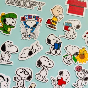 May include: A selection of Snoopy stickers depicting the character in various poses. The stickers show Snoopy in different outfits, holding items, and with text such as "Snoopy" and "Top Dog". The stickers are outlined in black and white with splashes of colour.