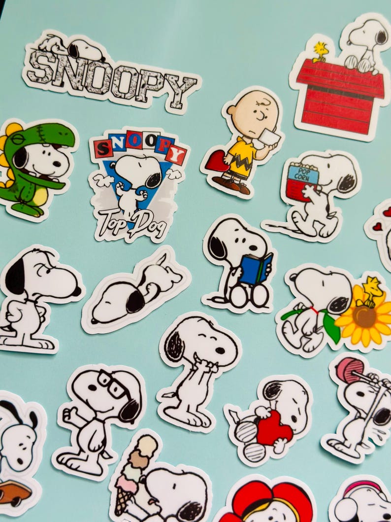 May include: A selection of Snoopy stickers depicting the character in various poses. The stickers show Snoopy in different outfits, holding items, and with text such as "Snoopy" and "Top Dog". The stickers are outlined in black and white with splashes of colour.